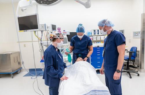Three clinic staff members in navy blue scrubs all standing around child patient laying down on on operating table.