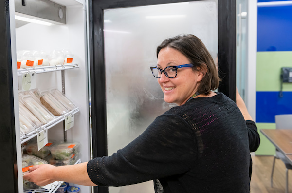 A smiling woman reaches into an open refrigerator in an office kitchen.