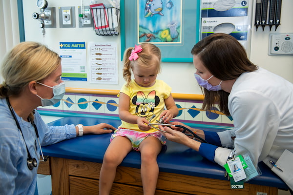 Two masked, female clinicians interact with a young girl on an exam table.