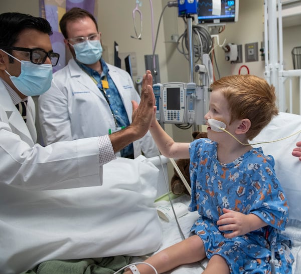 A physician high fives a young male child who is sitting in a hospital bed and has a feeding tube.