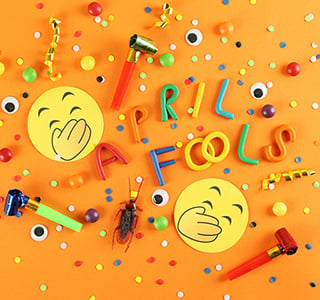 Confetti, laughing emojis, party horns and a fake bug spread out on a table with April Fools text.