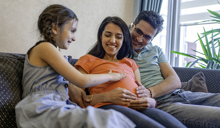 Young child touches mom's pregnant belly while sitting next to her and dad on the couch.