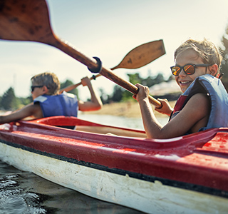 Two young boys in a canoe and wearing life jackets. They are using rowing paddles on a lake and smiling.