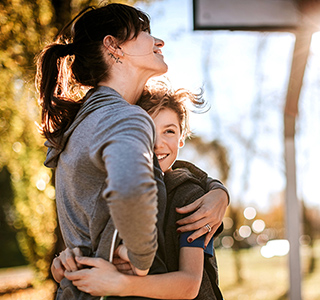 Mom hugs her preteen son. They are wearing sports clothes and near a basketball goal outside on a sunny day.