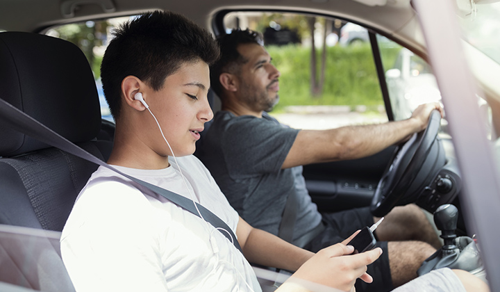 Teen boy sits in the passenger seat of the car with his dad listening to a phone with headphones.