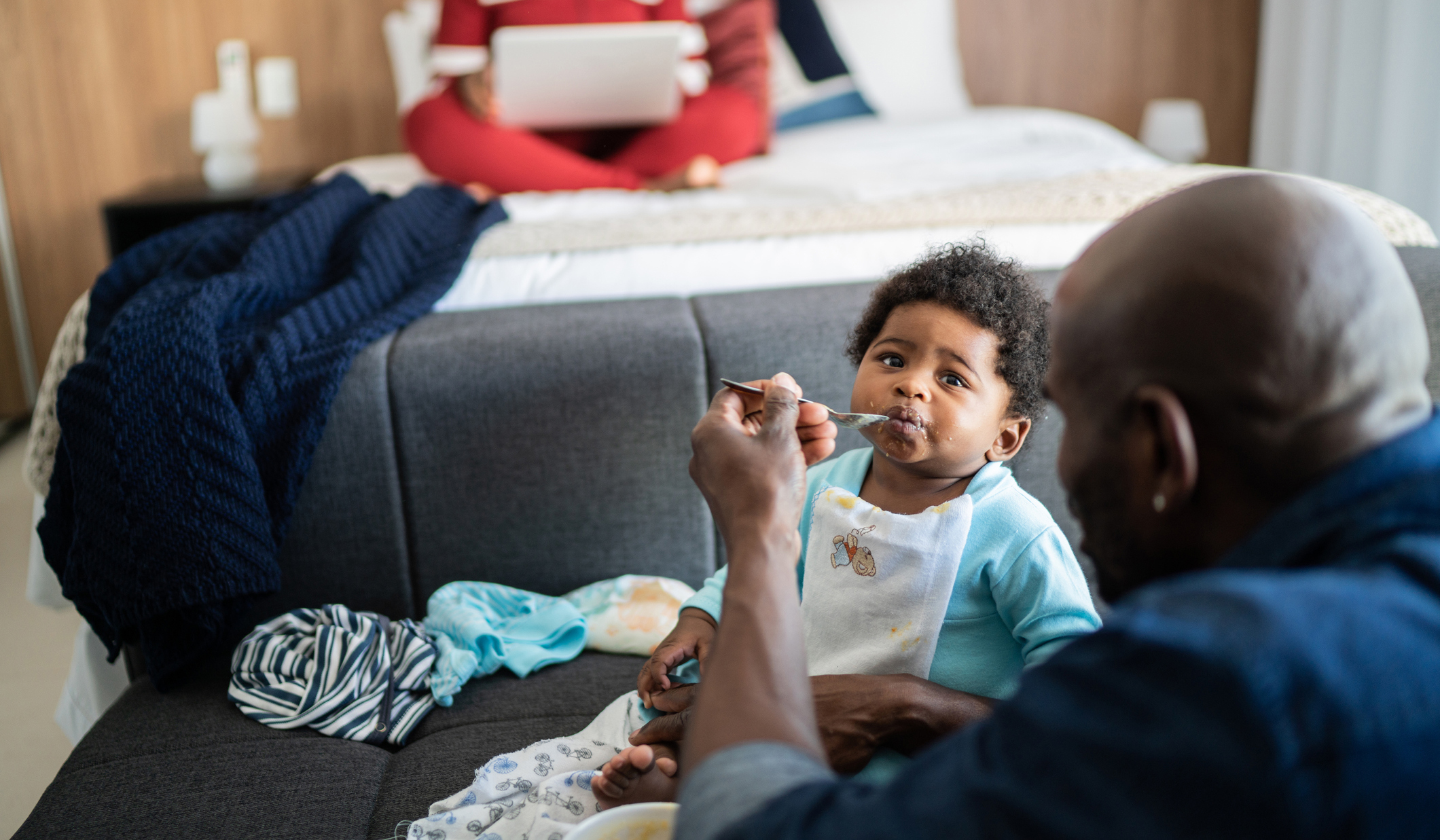 Dad is feeding baby some pureed food while mom sits on the bed in the background.