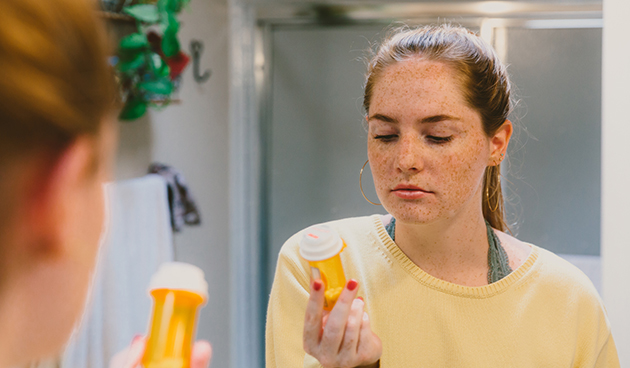 Young teen looks at a pill bottle while in front of a mirror.