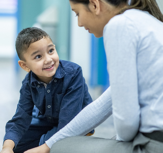 Young boy wearing a dark blue shirt sits on the floor next to a woman in a light blue shirt. They are playing with a toy on the ground.