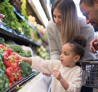 Young child reaching for red bell pepper in grocery store with parents behind her.