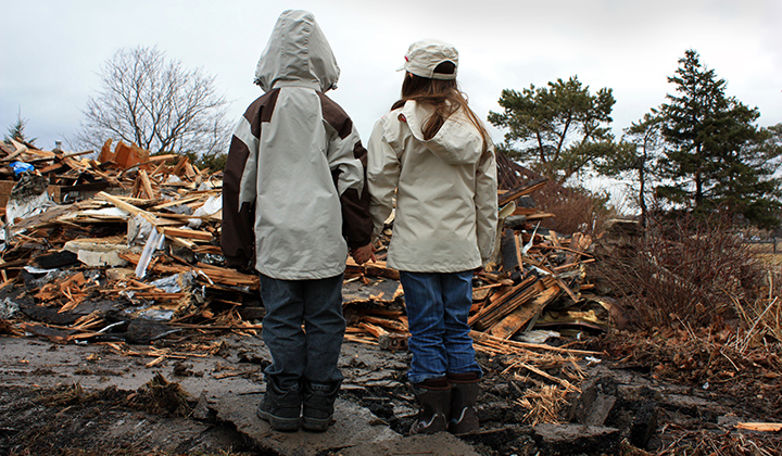 Two young children with backs to camera, hold hands and look at home debris after a storm. 