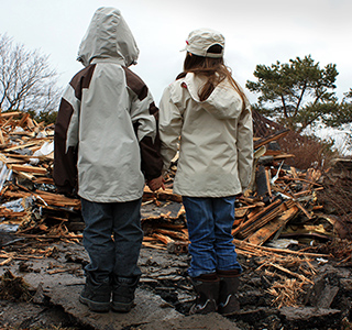 Two young children with backs to camera, hold hands and look at home debris after a storm. 