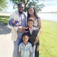 Mom and Dad posing outside by a body of water with their three children.