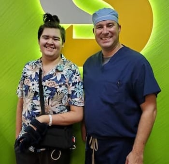 A teenage boy wearing a Hawaiian shirt and hospital bracelet stands next to a surgeon in blue scrubs.