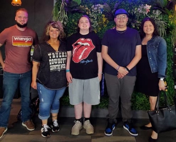 Five people stand in front of a wall of hanging plants. Two teenaged boys are in the middle with their moms on either side and one's older brother to the far left.