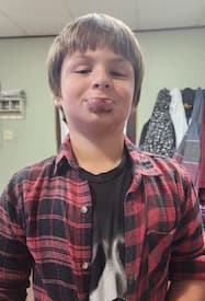 A boy with medium-length brown hair is wearing a red and black plaid shirt over a black tee shirt. His lower lip is visibly swollen after a medical procedure.