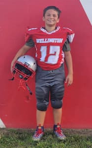 A middle school boy wears a football uniform with a red shirt and gray pants. His shirt has the team name “Wellington” and number 12. He is holding his helmet under his arm.