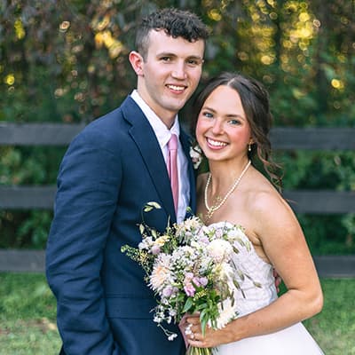 A young bride and groom smile, posing outside at their wedding in front of trees. Bride is wearing a strapless white wedding gown, holding a bouquet of flowers while the groom is in a navy blue suit with pink tie.