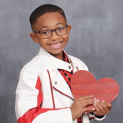 A boy in glasses poses in front of a photographer’s background. He's holding a wooden heart painted red and wearing a red and black checked shirt and white and red leather jacket.