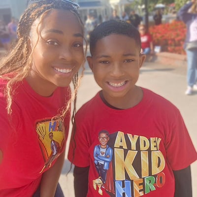 A mom and son smile at the camera wearing coordinated red t-shirts. We can see an amusement park street behind them. The boy’s t-shirt has a cartoon version of himself and lettering that reads “Ayden: Kid Hero.”
