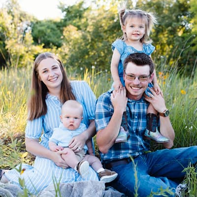 A family dressed in matching blue shades sits in a field of grass. On the left, the mother holds a baby boy while his toddler sister sits on the dad’s shoulder to their right.