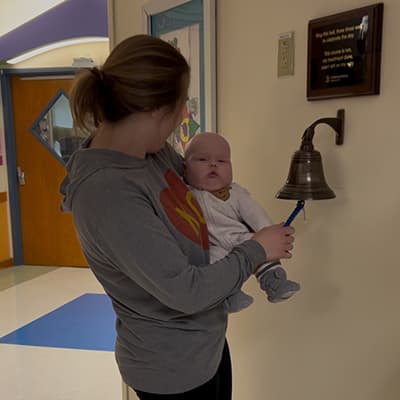 Mother carrying baby patient in hospital hallway wearing gray KC pullover.