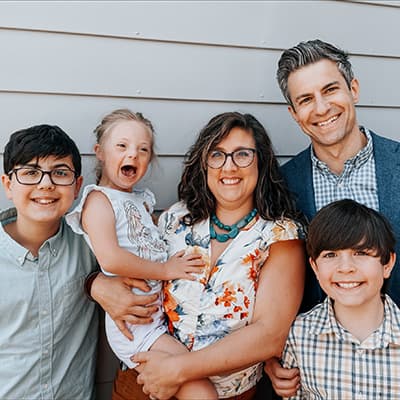 A family of five stands together in front of a light-colored exterior wall, with one adult holding a young child.