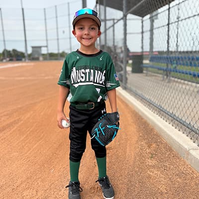 A young boy on a baseball field poses with a glove and ball. He is wearing a green jersey with “Mustangs” on it and baseball pants, cleats and cap.