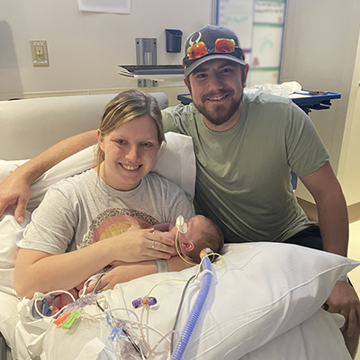 A mother holds a small baby in a hospital room while the father sits to the side with his arm around them. The baby is cradled on pillows and has medical tubing coming from her tiny body. 