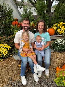 Four people sit together on hay bales outdoors, surrounded by colorful fall flowers and pumpkins.