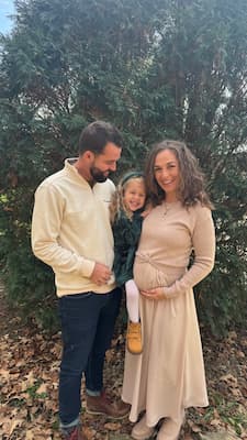Three people stand outdoors in front of evergreen trees, with one adult resting a hand on a visibly pregnant belly and a child standing between the two adults.