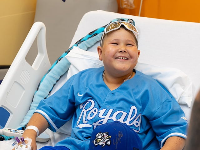 Jaxson sits in a hospital bed wearing a Kansas City Royals jersey and holding a signed baseball cap during a visit at Children's Mercy. 