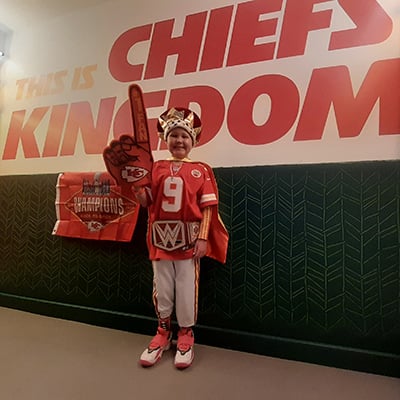  Jaxson stands in front of a large “This Is Chiefs Kingdom” wall graphic wearing Kansas City Chiefs apparel and holding a foam finger.