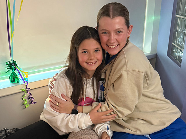 Jenna and Liv sit together on a hospital bed and pose for a photo. Liv has one arm around Jenna. A large plush pillow sits in the foreground and colorful decorations hang near a window behind them.
