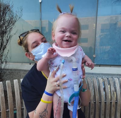  An adult holds a child with a tracheostomy outdoors near a bench and building windows.