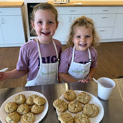 Two little girls, around 5 and 3, stand in front of two paper plates of cookies on a stainless steel countertop. They are in a large kitchen space and are wearing matching purple t-shirts and white aprons that read “TasteBuds Kitchen”.