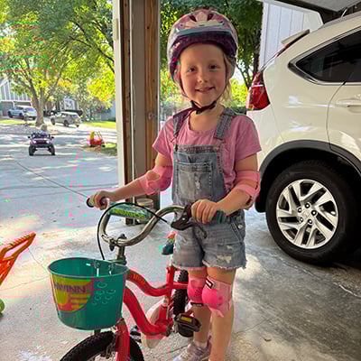 A young girl poses in a garage with a brightly colored bike. She is wearing a bike helmet, pink elbow and knee pads and overall shorts.