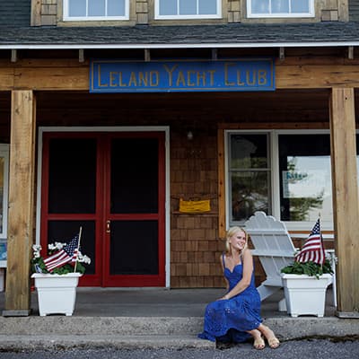 A smiling young woman in a blue dress sits on the front porch of a rustic two-story building covered in shaker shingles. A blue hand-carved sign hanging above the red front door reads "Leland Yacht Club.” Two white planters on the patio hold American flags, and there’s a white deck chair behind the young woman.