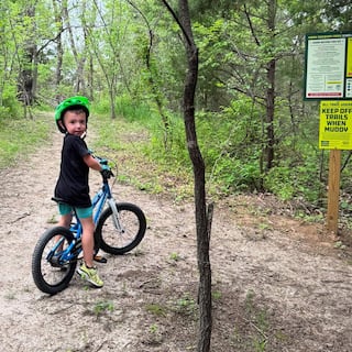 Koda wearing a bright green helmet standing with a blue bicycle on a wooded trail, looking back toward the camera near a posted trail information sign.