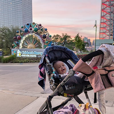 Lilly sits in a stroller outside Children's Mercy Adele Hall Campus with hospital signage visible in the background.