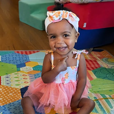 Lilly sits on a colorful play mat wearing a patterned dress and headband.