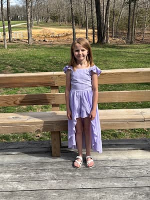 A young girl wearing a checked purple dress and white sandals poses on a deck. There’s a big yard with leafless trees and a long driveway behind her. 