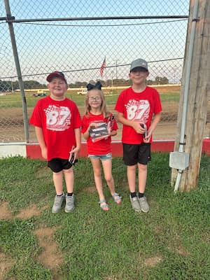 Three kids in matching red t-shirts that read “Team 87 Motorsports” smile in front of a dirt racetrack with an American flag in the background. 