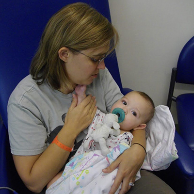 A mom wearing an orange hospital bracelet holds a baby wrapped in a blanket and using a pacifier. They are sitting on a blue chair in what looks like a waiting room. 