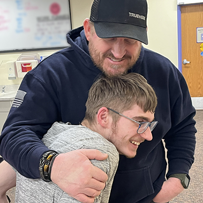 A dad in a navy blue sweatshirt and black ball cap wraps one arm around his teen son in a proud hug. Both dad and son have light brown hair and beards. The son is smiling and returning the hug.
