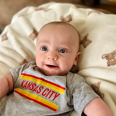 A baby with big blue eyes smiles at the camera. He is wearing a onesie that reads “Kansas City” and is laying on a cream blanket with teddy bear illustrations. 