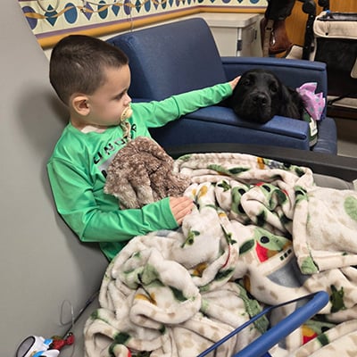 Child sitting in a hospital chair with a blanket and a black dog resting nearby