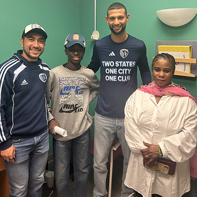 A patient stands in a clinic exam room with several visitors wearing soccer apparel, positioned together for a photo during a hospital visit.