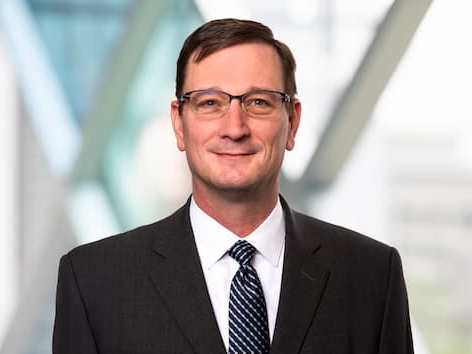 Professional headshot of a person wearing glasses, a dark suit, white shirt, and striped tie, standing in front of a softly blurred architectural background with large geometric window structures.
