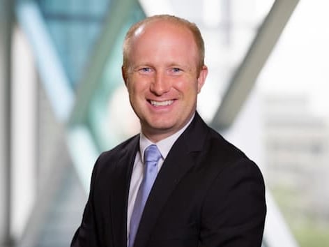 Professional headshot of a person wearing a dark suit jacket, light blue shirt, and blue tie, seated in front of a softly blurred office background with geometric glass and metal architecture.