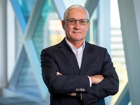 Professional headshot of a person wearing glasses and a dark blazer, standing with arms crossed in a modern building with large geometric windows and blurred architectural elements in the background.